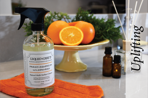 Image of the Fresh-Squeezed Wilderness Natural Multi-Surface Cleaner with a stand of oranges and essential oil bottles in the background. The bottle is on top of an orange microfibre cloth. There is cedarwood, orange, and clary sage essential oils infused in the cleaner. The word uplifted in the image depicts the mood intended by the cleaner.